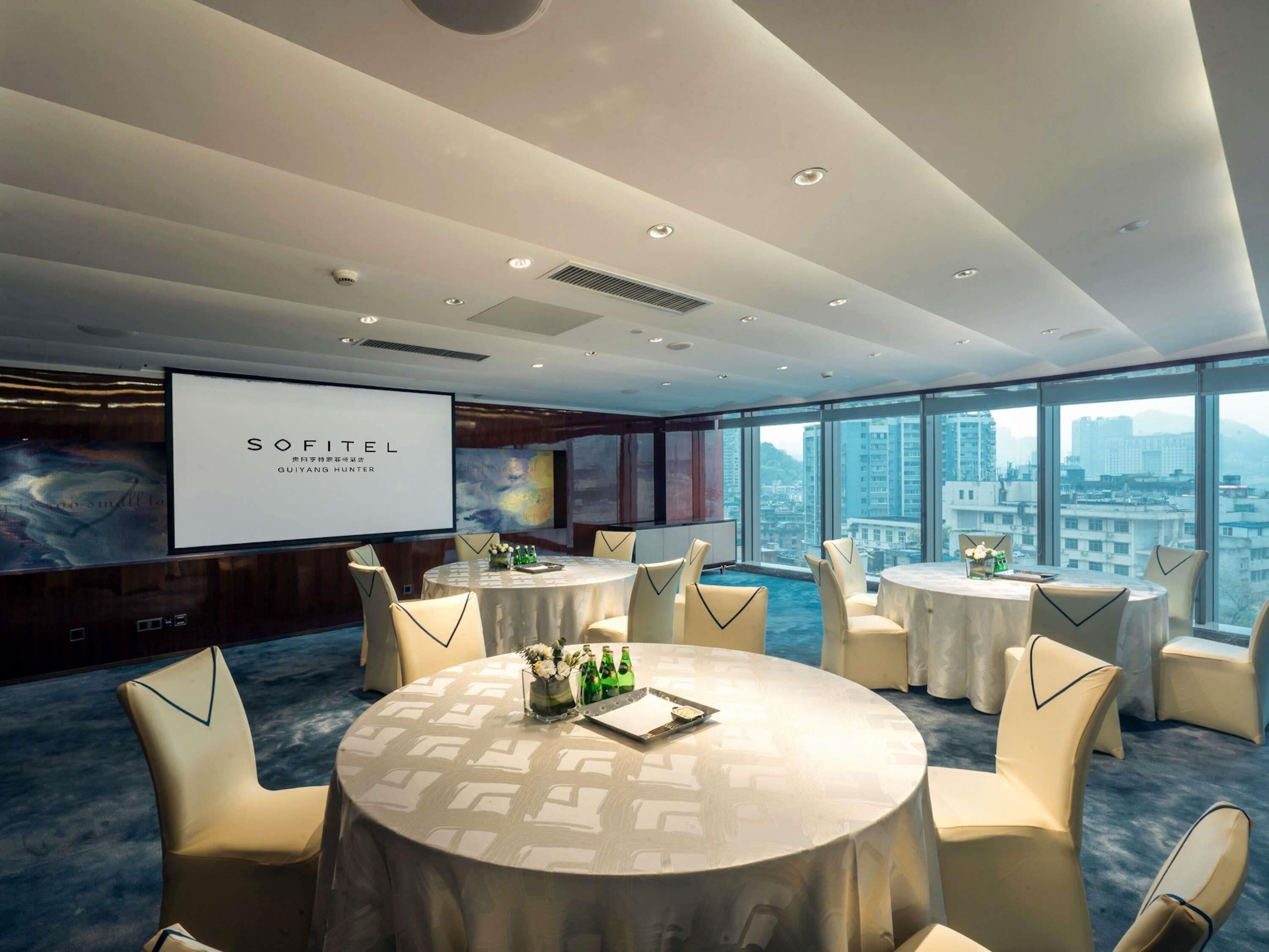 Un salón de conferencias con una pantalla grande que muestra 'SOFITEL GUYANG HUNTER', mesas redondas con sillas y vistas a la ciudad a través de grandes ventanales.
