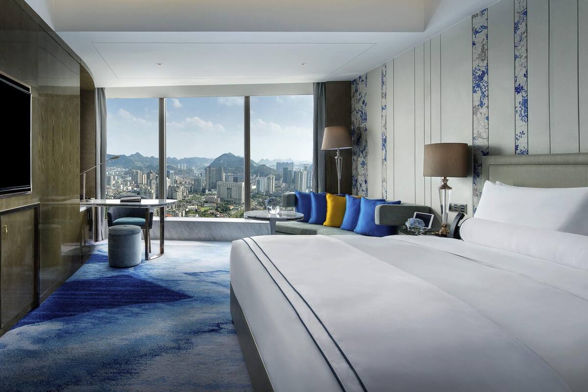 A hotel room with a large bed, a desk, a chair, and a view of a cityscape.