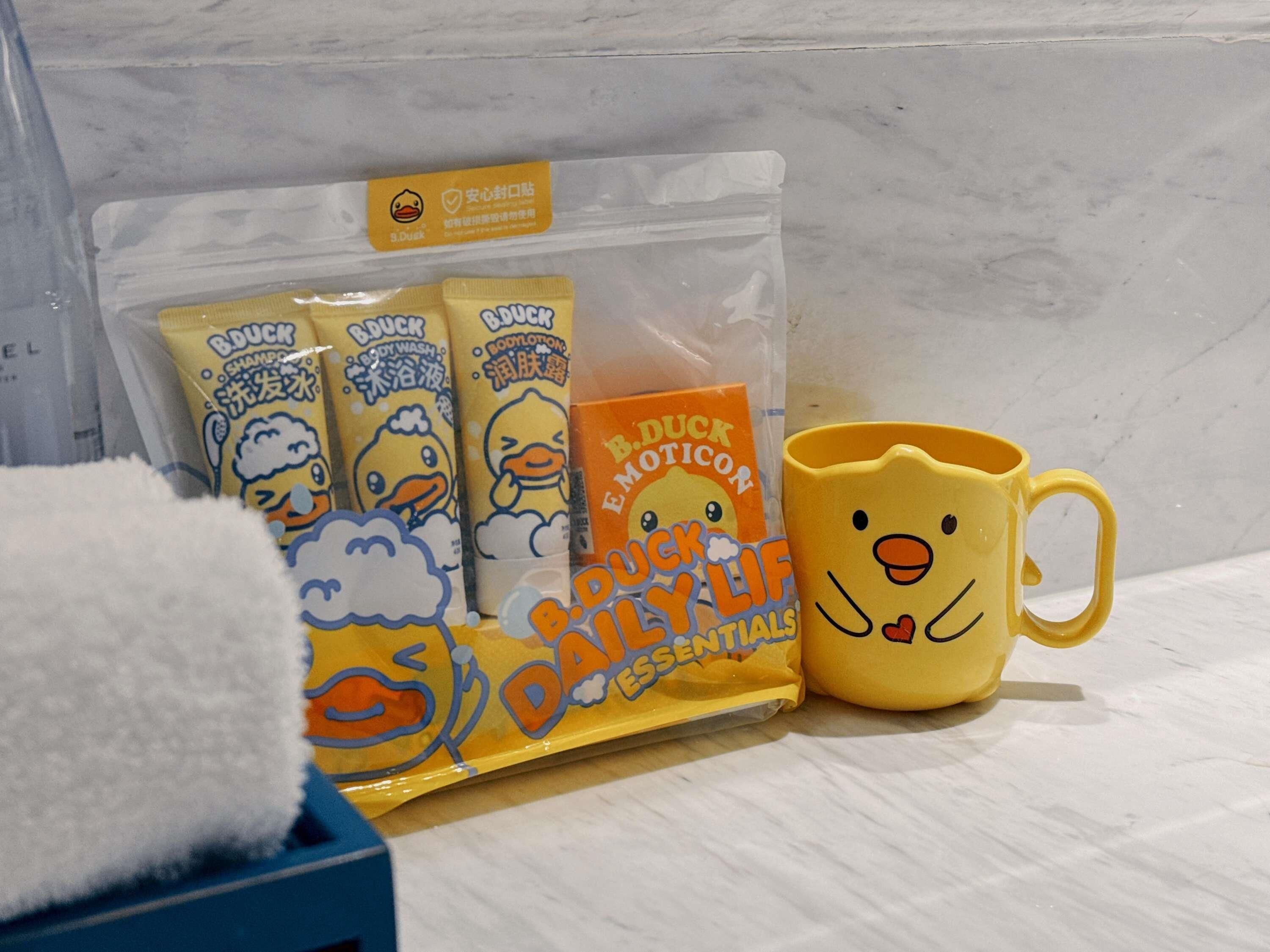A bathroom with a white towel, a yellow mug with a duck design, and a package of duck-themed toiletries.