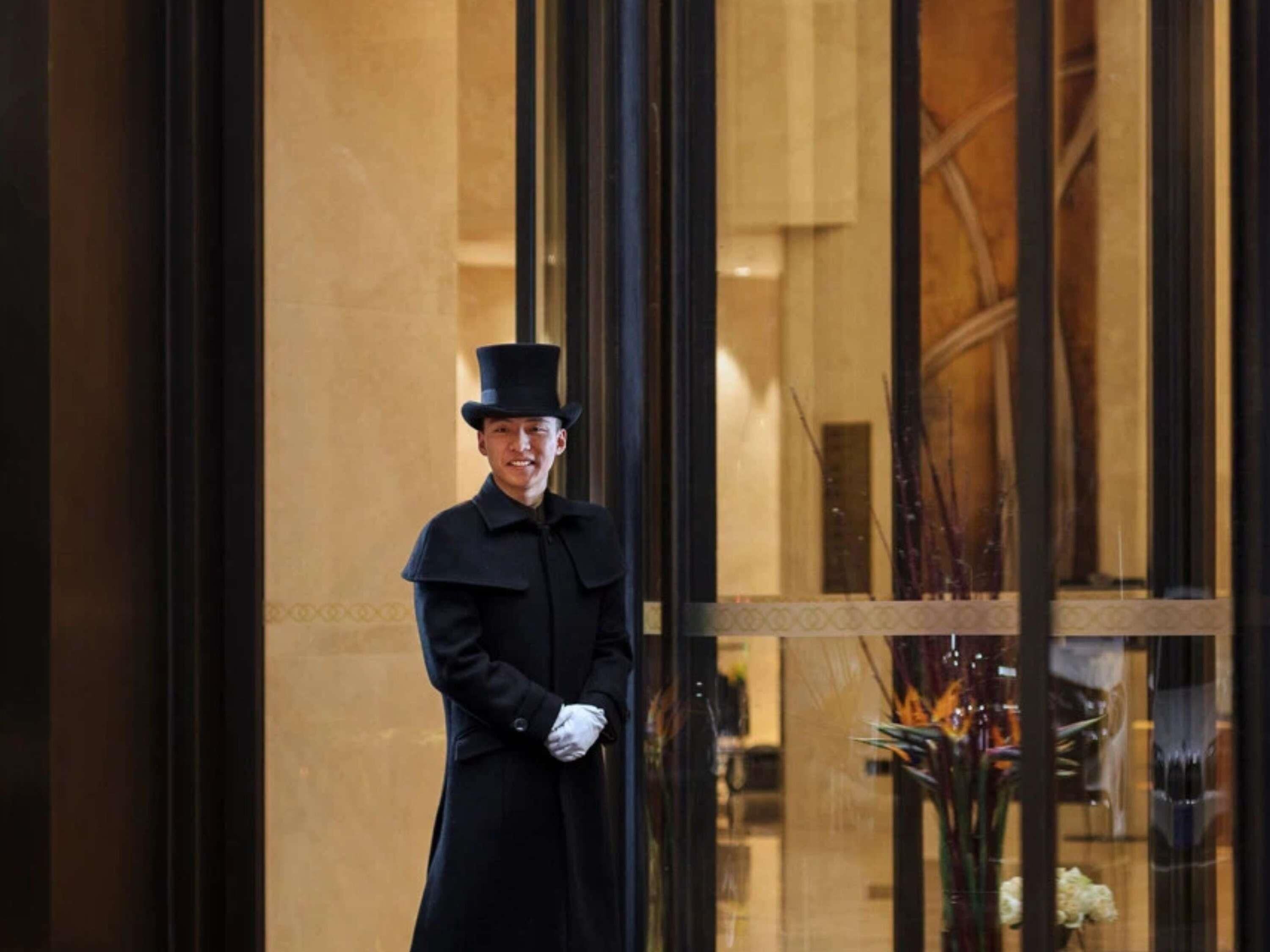 A person in a black coat and top hat standing in front of a building with large glass windows.