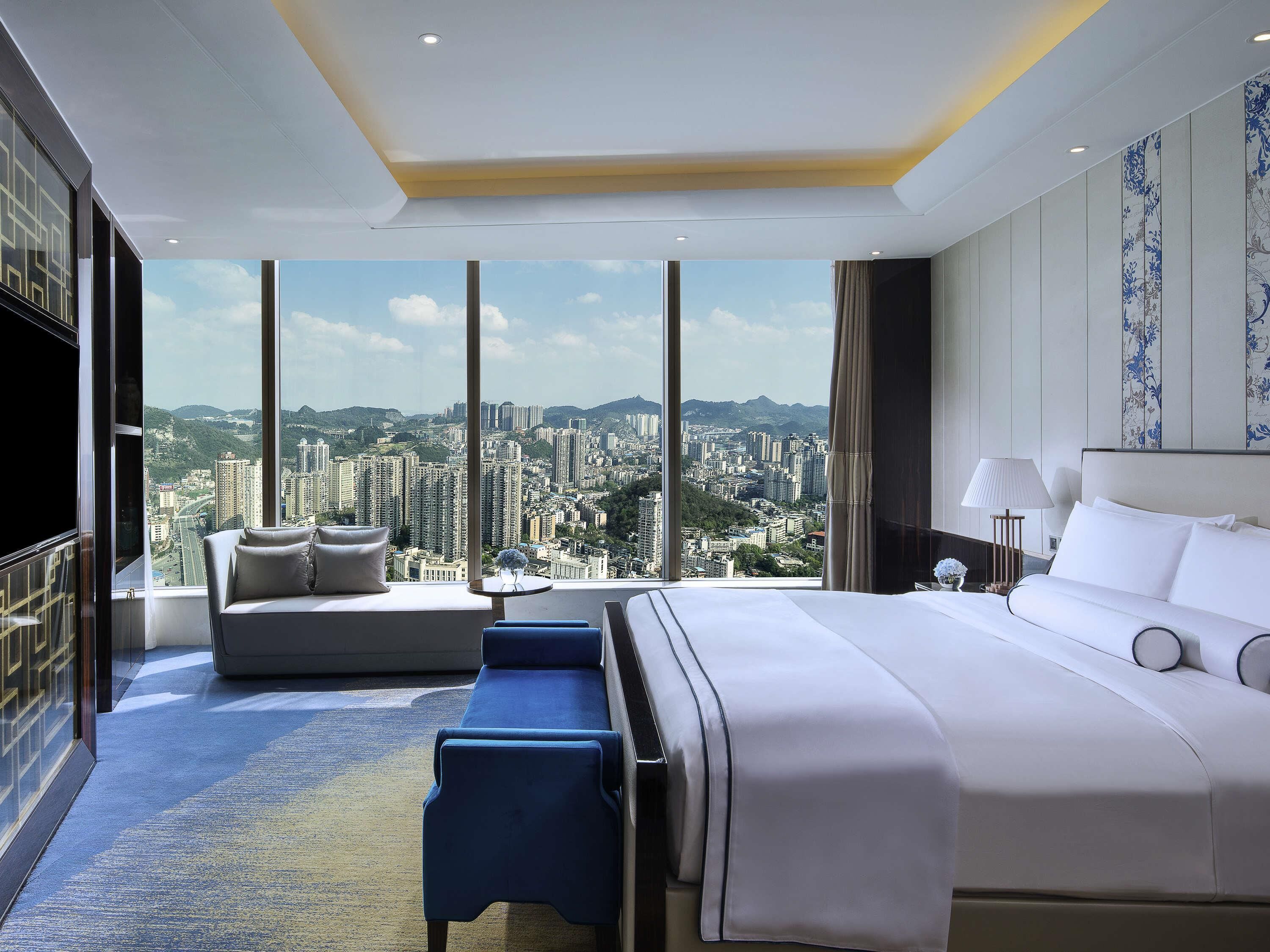 Ein Hotelzimmer mit einem großen Bett, einer Couch, einem kleinen Tisch und Blick auf eine Stadtlandschaft.