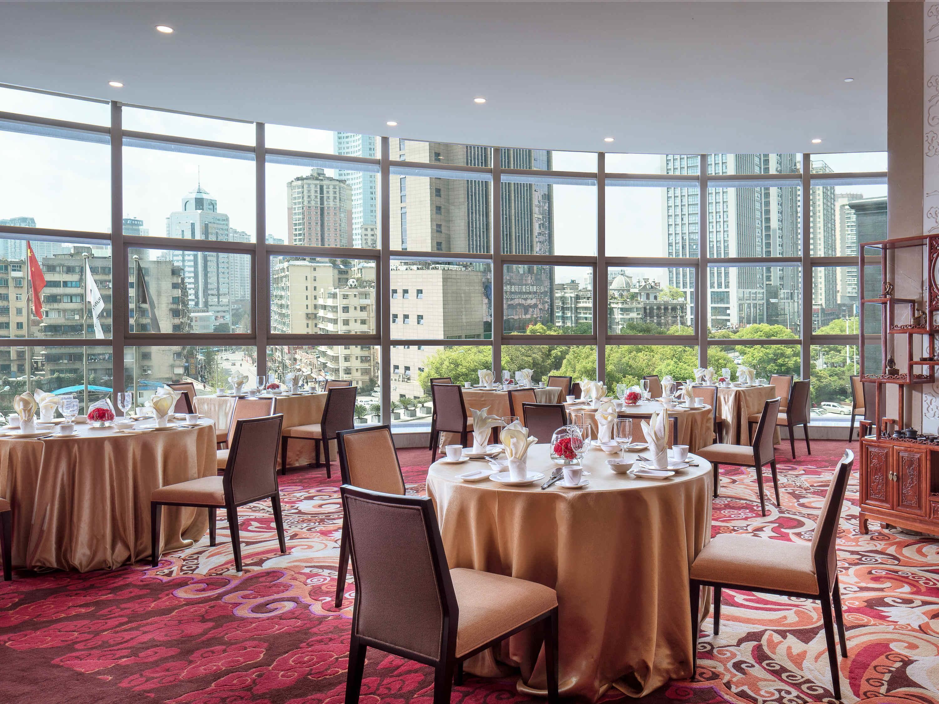 A dining area with round tables set for service, large windows offering a city view, and a patterned carpet.