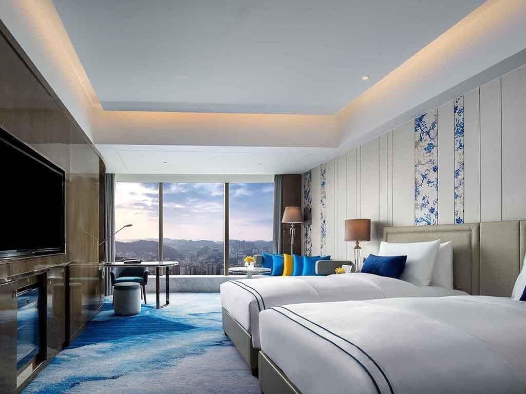 A modern hotel room with a large bed, a TV, a desk, and a view of the city.