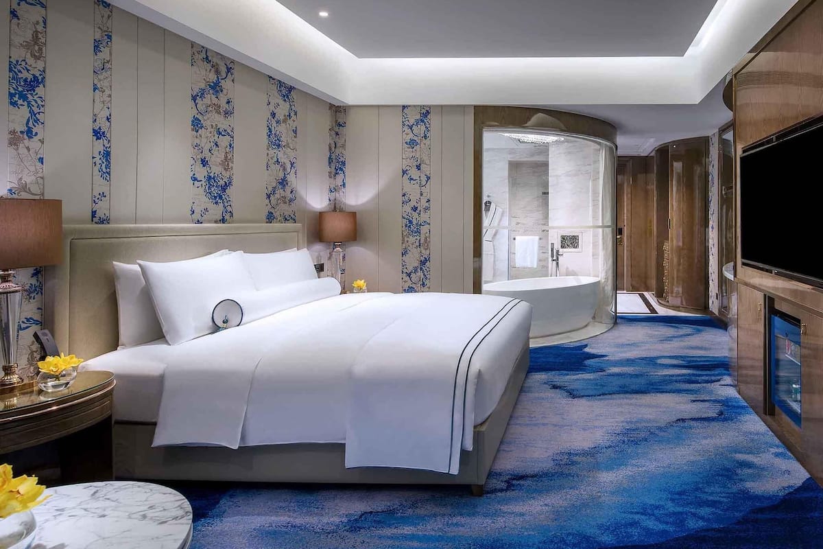 Une chambre d’hôtel avec un grand lit, une table ronde ornée d’un vase de fleurs, une table de chevet, une lampe et un grand miroir.