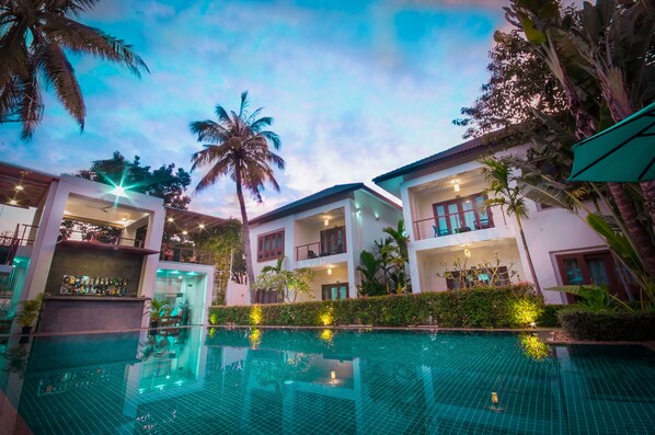Vista de la piscina de un resort con varias construcciones, palmeras y cielo despejado.