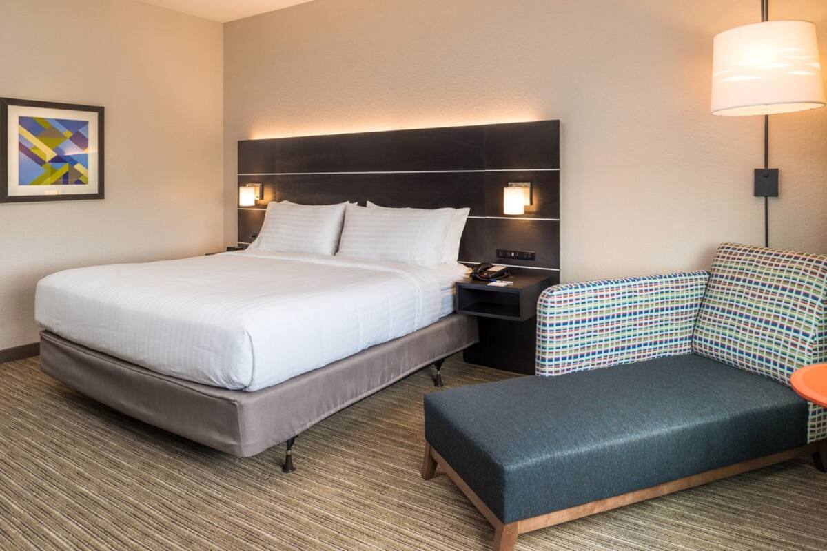 Una habitación de hotel con una cama grande, una mesita de noche, una lámpara y un sillón estampado.