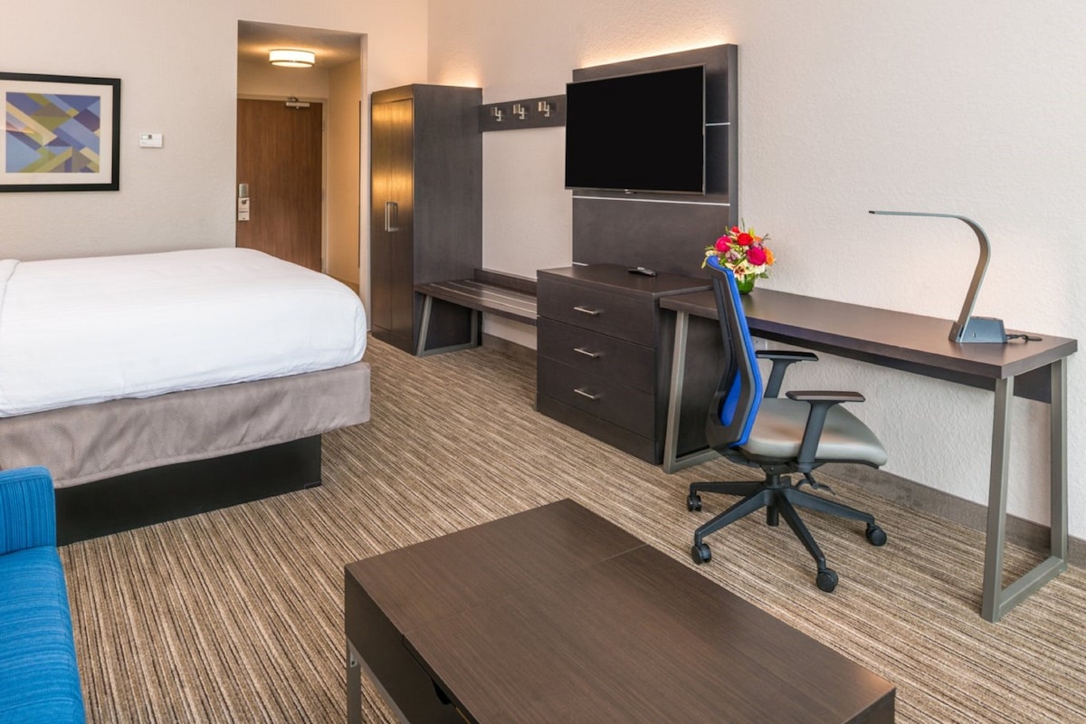 A hotel room with a bed, a desk with a chair, a TV, and a small table.
