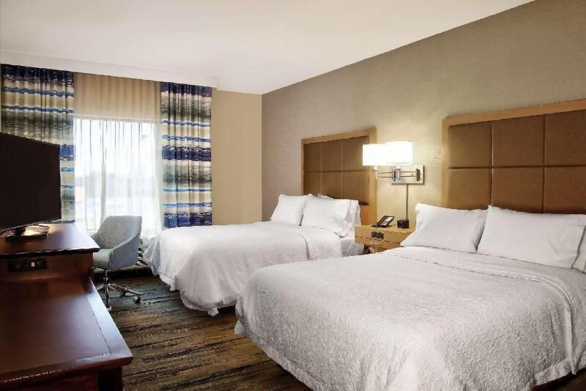 A hotel room with two beds, a desk, a chair, a television, and a window with curtains.