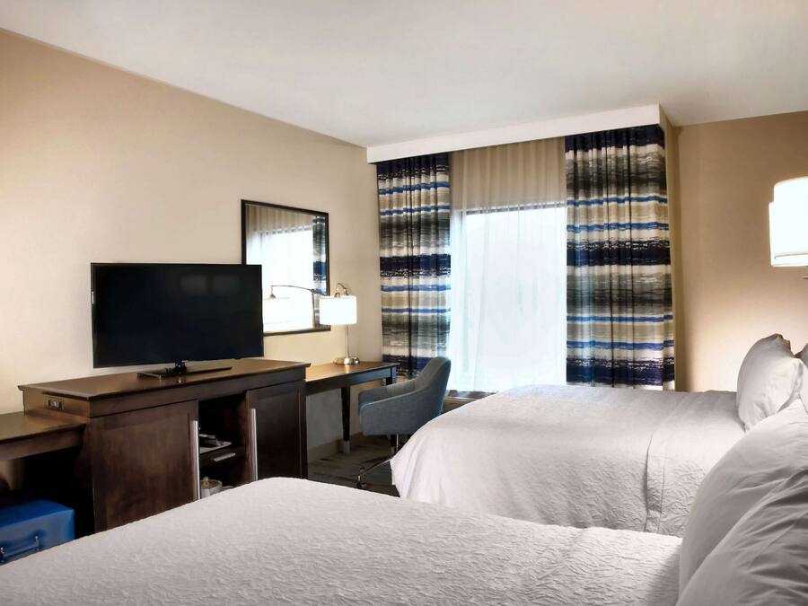 A hotel room with two beds, a desk with a TV, a chair, and a large window with striped curtains.