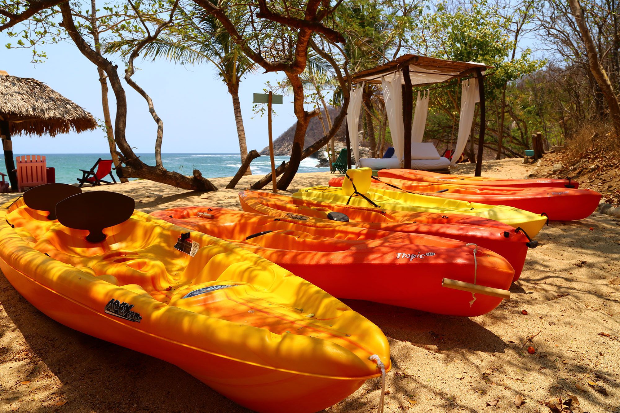Una playa con varios kayaks amarillos y rojos alineados en la arena, una sombrilla de paja y un pequeño pabellón.