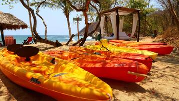 Una playa con varios kayaks amarillos y rojos alineados en la arena, una sombrilla de paja y un pequeño pabellón.