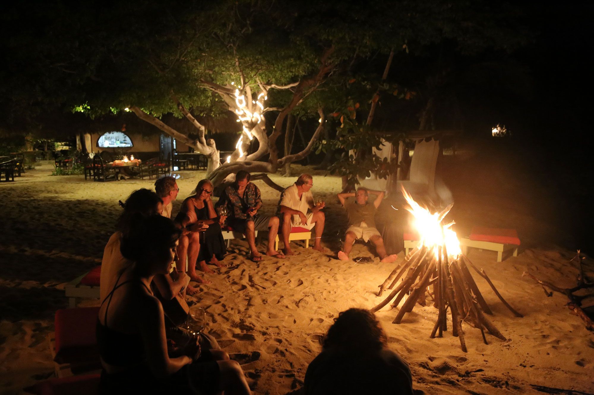 A group of people sitting around a campfire on a sandy beach at night.