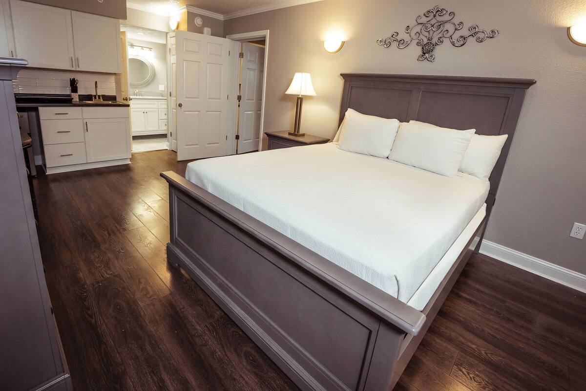 A bedroom with a large bed, a nightstand with a lamp, a dresser, and a walk-in closet.