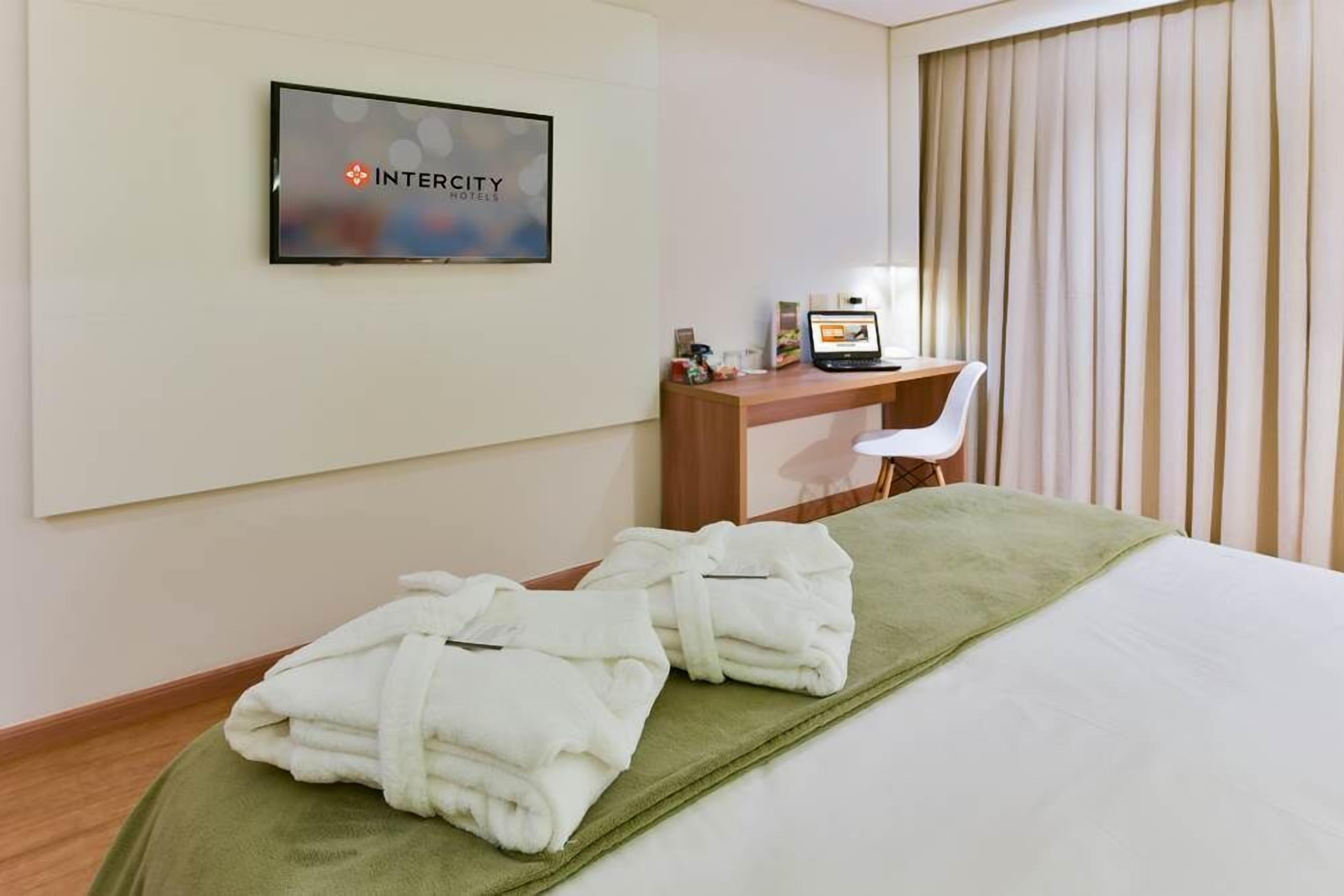 A hotel room with a bed, a desk with a computer, and a TV displaying the hotel name.