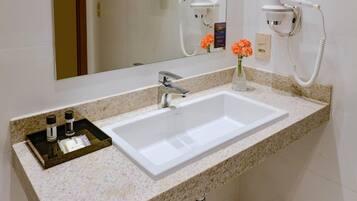 Een badkamer met een witte wastafel, een granieten aanrechtblad en een spiegel. Twee haardrogers zijn aan de muur bevestigd en er staat een vaas met bloemen op het aanrecht.