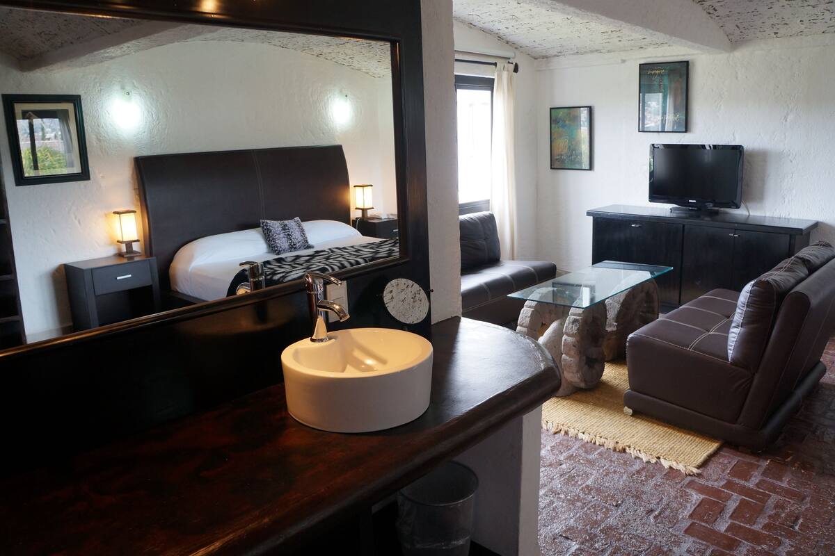 Habitación de hotel con cama, televisión, sofá, mesa de centro y baño con lavamanos y espejo.