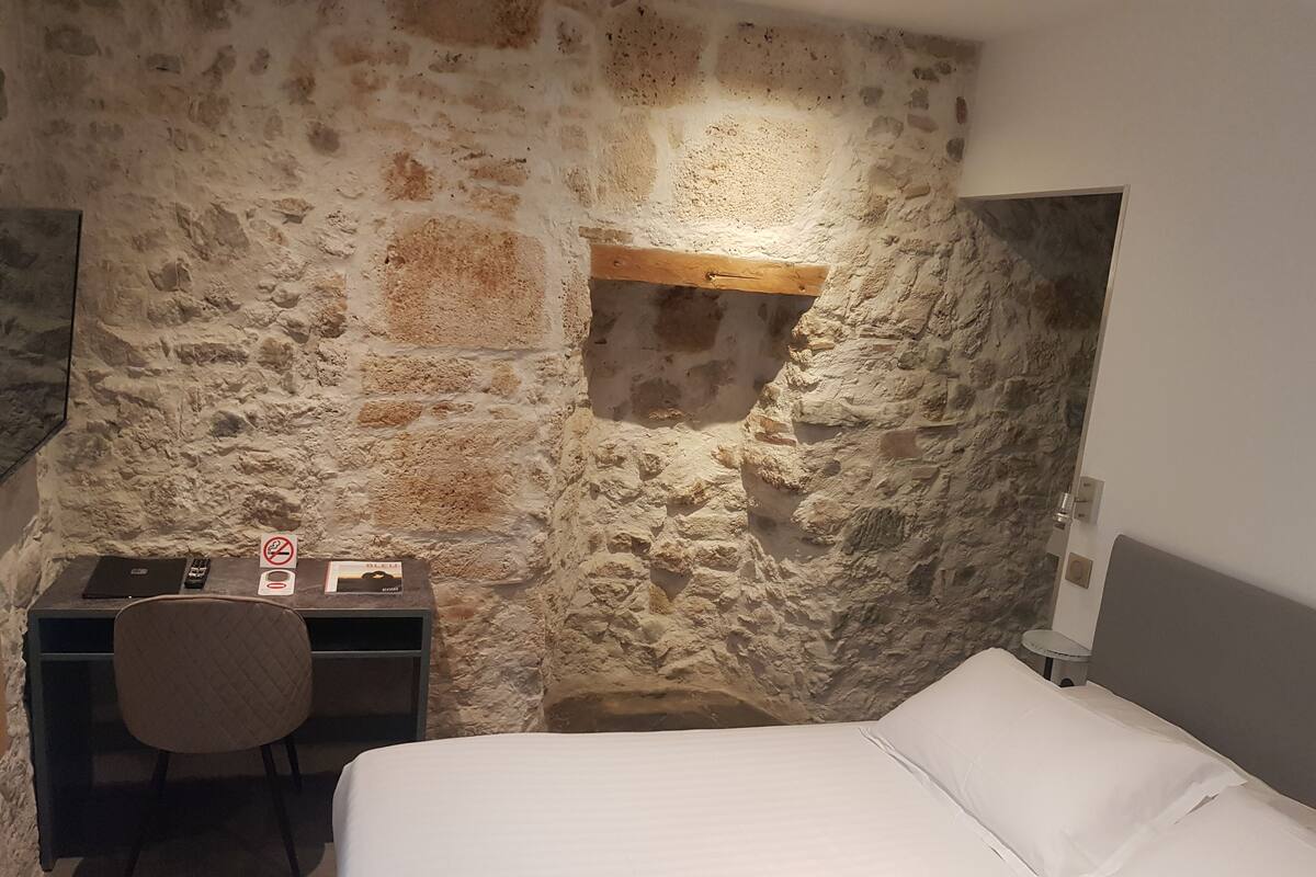 Quarto com parede de pedra, uma cama, uma escrivaninha com cadeira e uma pequena estante.
