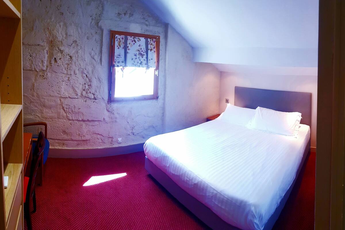 Quarto de hotel com cama, janela com cortinas florais, área de chuveiro e vaso sanitário.