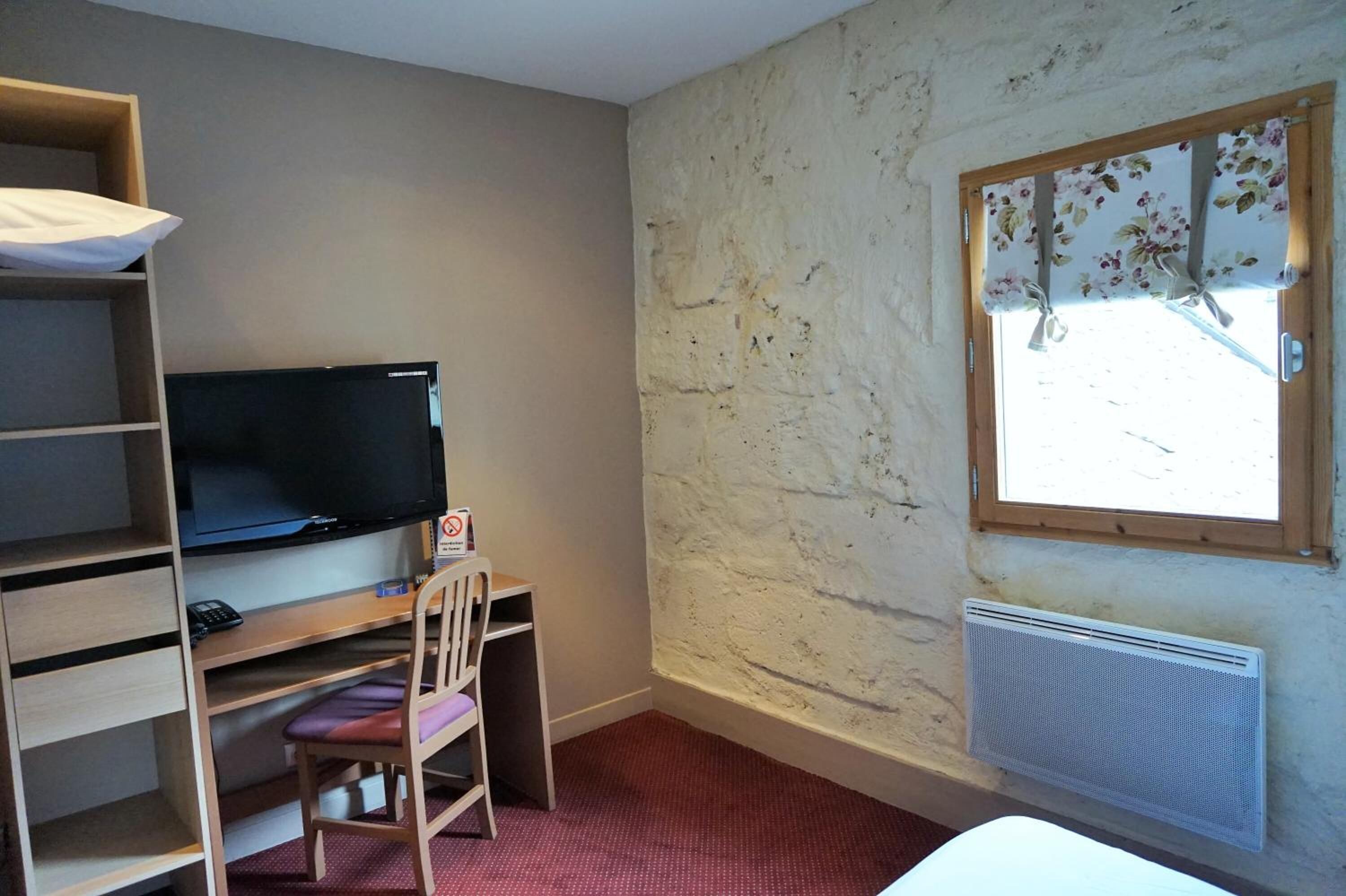 Une pièce avec un téléviseur posé sur une étagère en bois, une chaise, un radiateur et une fenêtre ornée d’un rideau à motifs floraux.