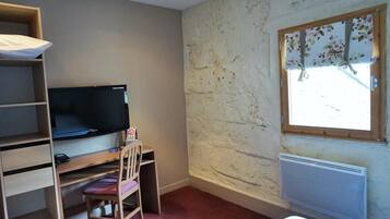 Une pièce avec un téléviseur posé sur une étagère en bois, une chaise, un radiateur et une fenêtre ornée d’un rideau à motifs floraux.