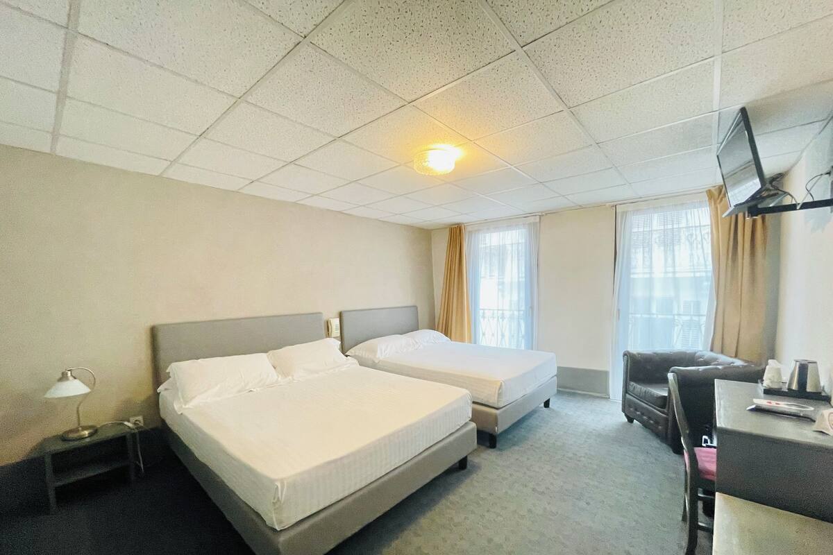 Quarto de hotel com duas camas, uma escrivaninha com cadeira, televisão e janela com cortinas.