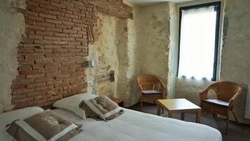 A room with a brick wall, a bed with pillows, a small table, two wicker chairs, and a window with a view.