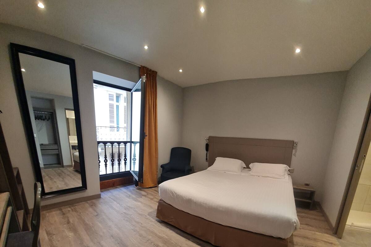 Quarto de hotel com cama grande, cadeira, escrivaninha, espelho e vista para o exterior.