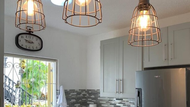 Una cocina moderna con un refrigerador de acero inoxidable, un horno empotrado y una placa de inducción negra. Se observan tres lámparas colgantes de diseño entramado, y un reloj en la pared.