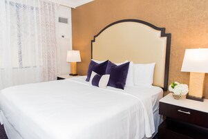 Una cama bien hecha con almohadas blancas y moradas, un cabecero de madera y dos lámparas de noche.