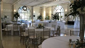 A spacious hall with multiple round tables and chairs, large arched windows, and decorative floral arrangements.