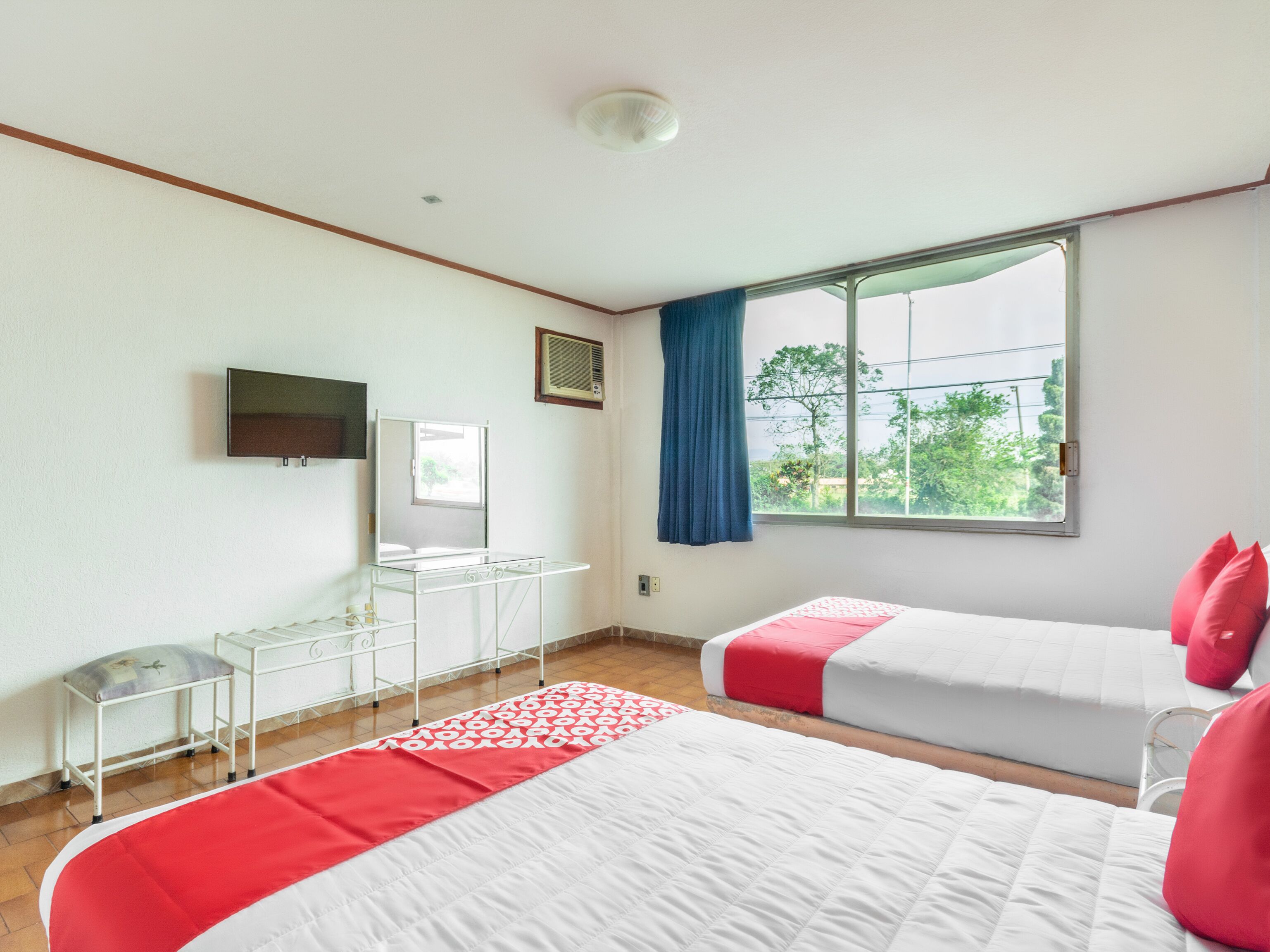 A hotel room with two beds, a TV, a desk, and a window with a view of trees.