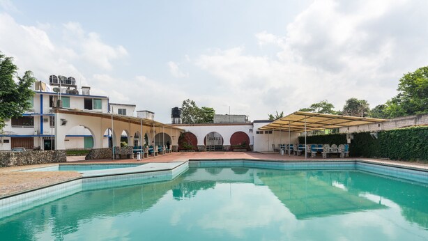 Una piscina con borde de piedra, una zona de estar cubierta y un edificio con varios arcos al fondo.