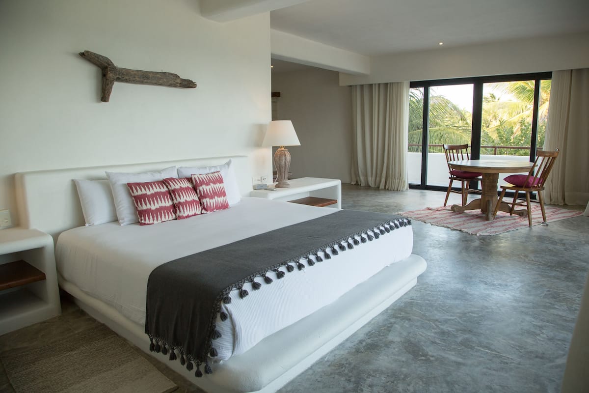 A bedroom with a large bed, bedside tables, a lamp, a dining area with chairs, and a view through large windows.