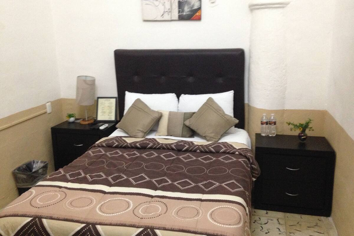 Uma cama bem arrumada com colcha estampada, cabeceira preta e duas mesas de cabeceira com abajures e um quadro emoldurado.