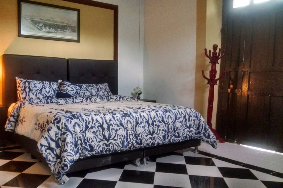 Um quarto com chão xadrez, uma cama com colcha estampada, um criado-mudo com abajur e uma porta com puxador decorativo.