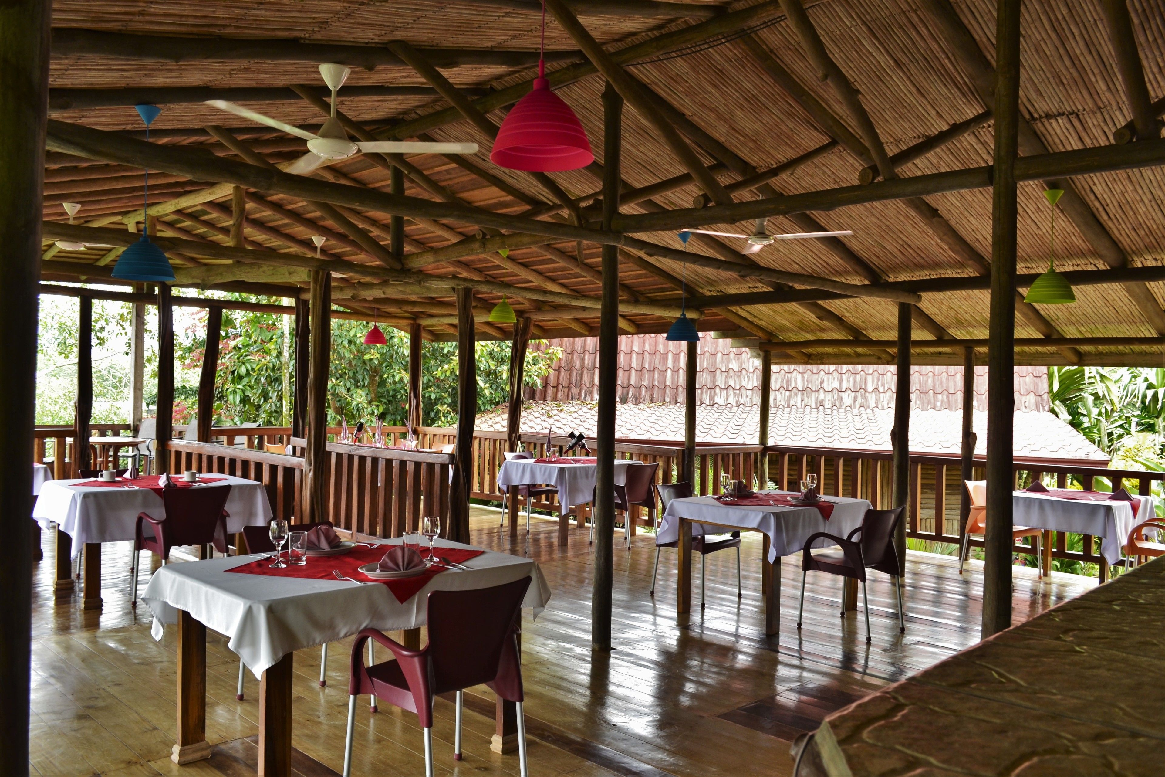 An outdoor restaurant with wooden tables and chairs, covered by a thatched roof, and surrounded by greenery.