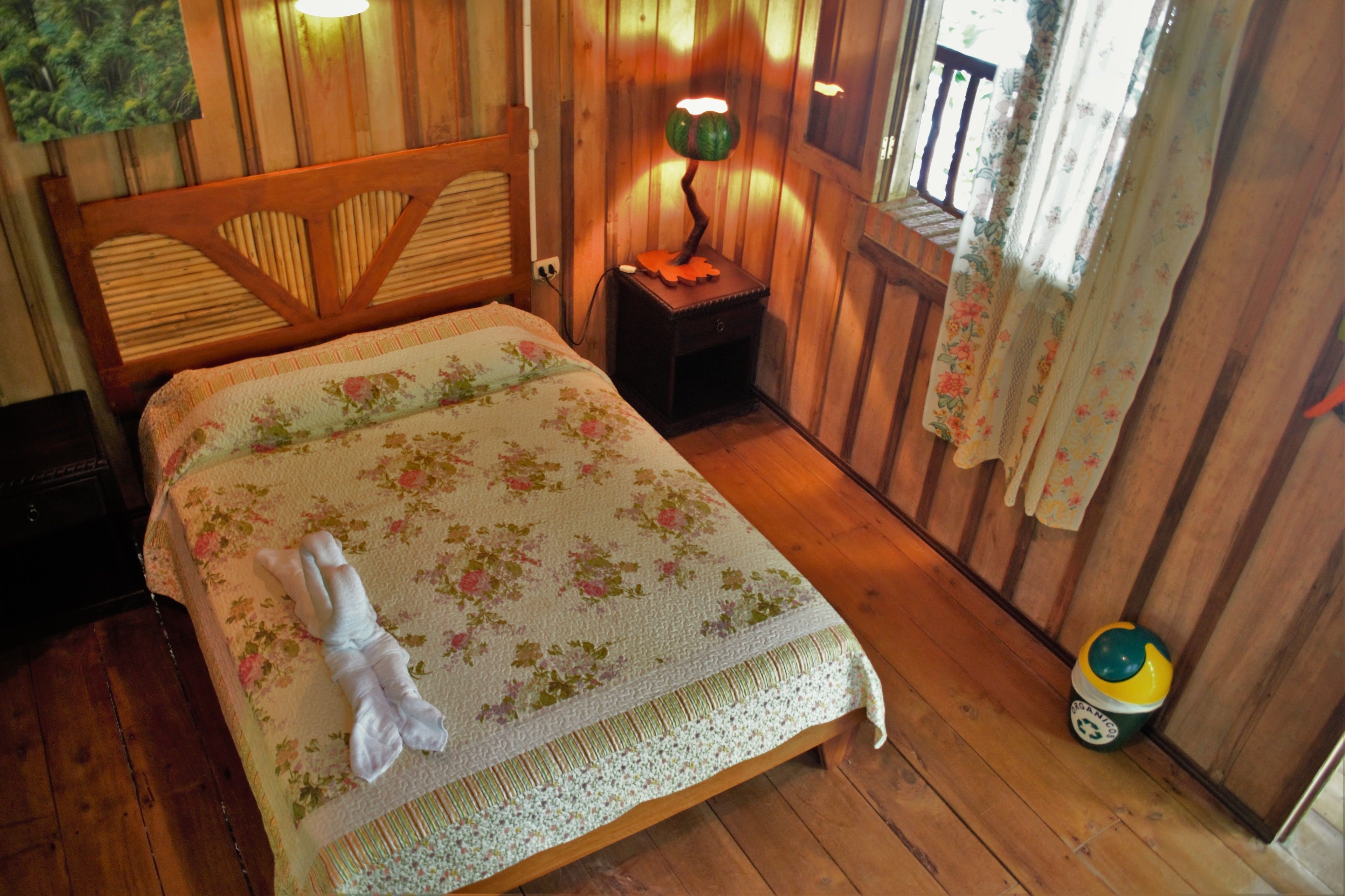 A wooden room with a bed, a nightstand, a lamp, and a small trash bin.