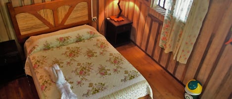 A wooden room with a bed, a nightstand, a lamp, and a small trash bin.