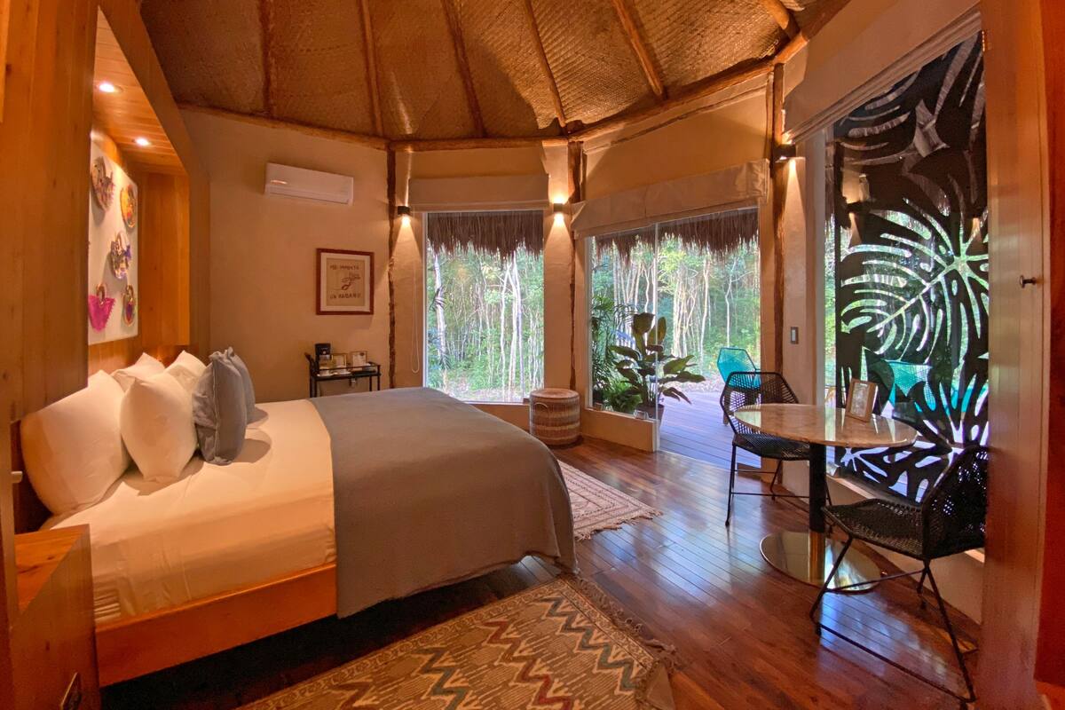 Quarto com cama, mesa e cadeira. Janela com vista para árvores.