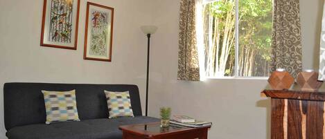 A living room with a dark grey sofa, two framed wall art pieces, a floor lamp, a window with patterned curtains, and a small wooden table with books and a plant.