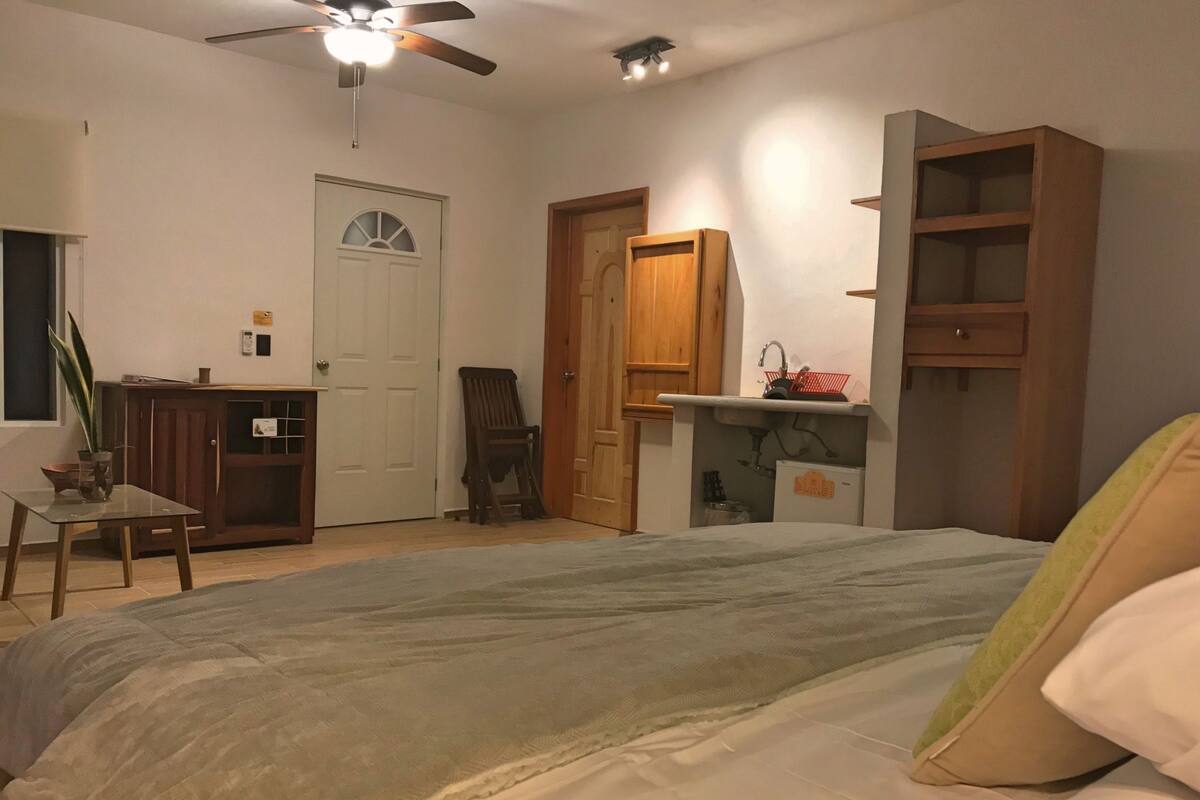 Um quarto com uma cama, uma escrivaninha, uma cadeira, um ventilador de teto e uma pequena cozinha compacta.