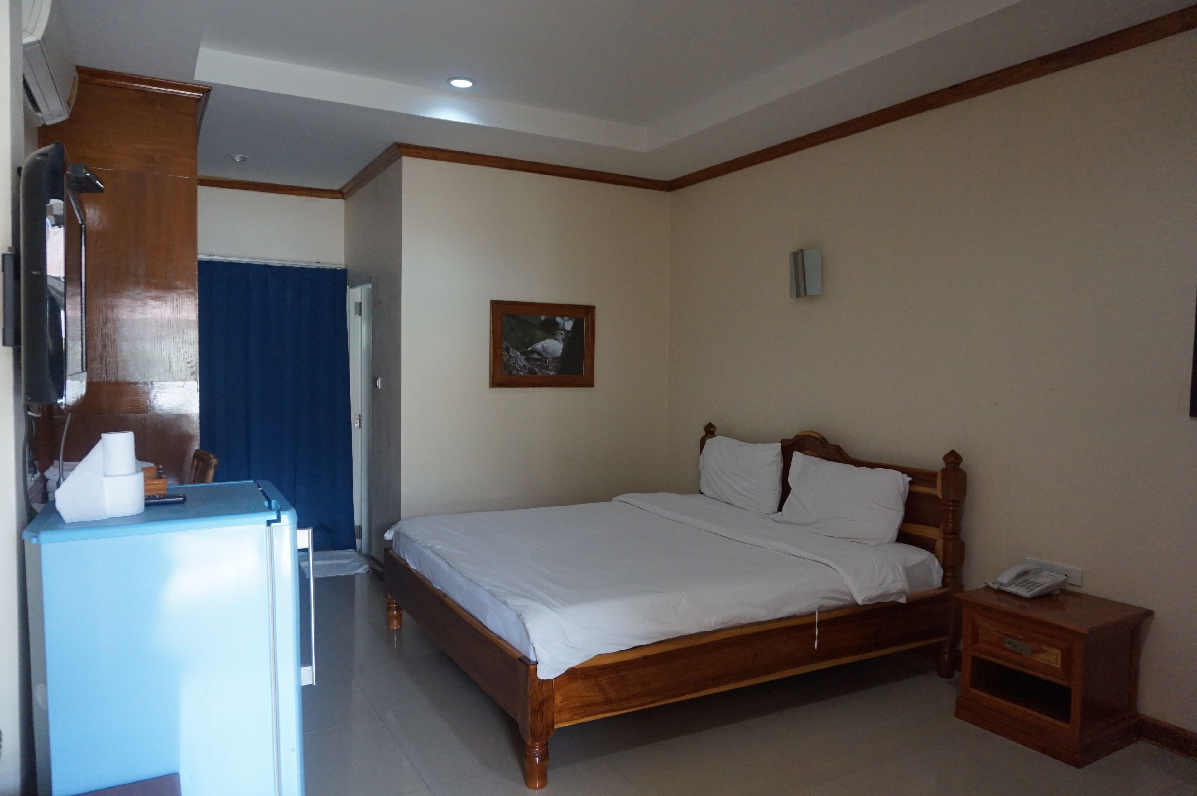 A bedroom with a wooden bed, a small bedside table with a phone, a blue curtain, a wall-mounted picture, and a small desk with a lamp.