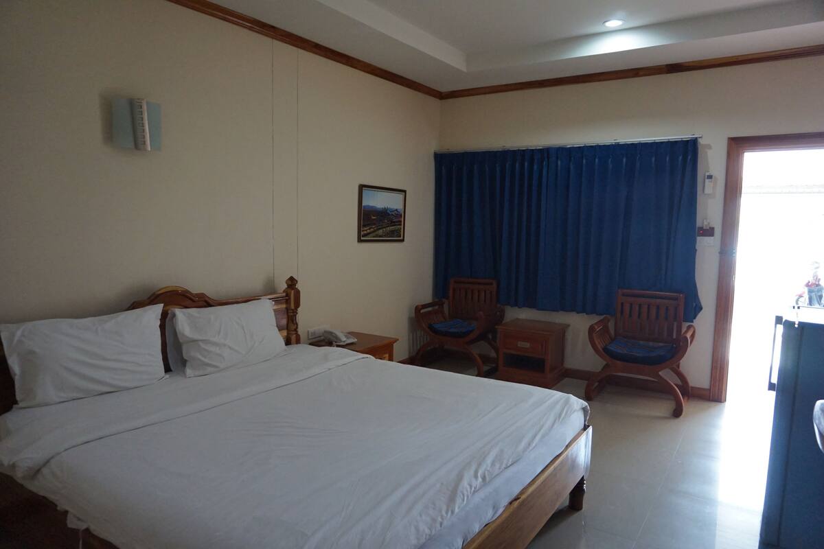 Habitación de hotel con una cama grande, dos sillas de madera, una mesita y una ventana con cortinas azules.