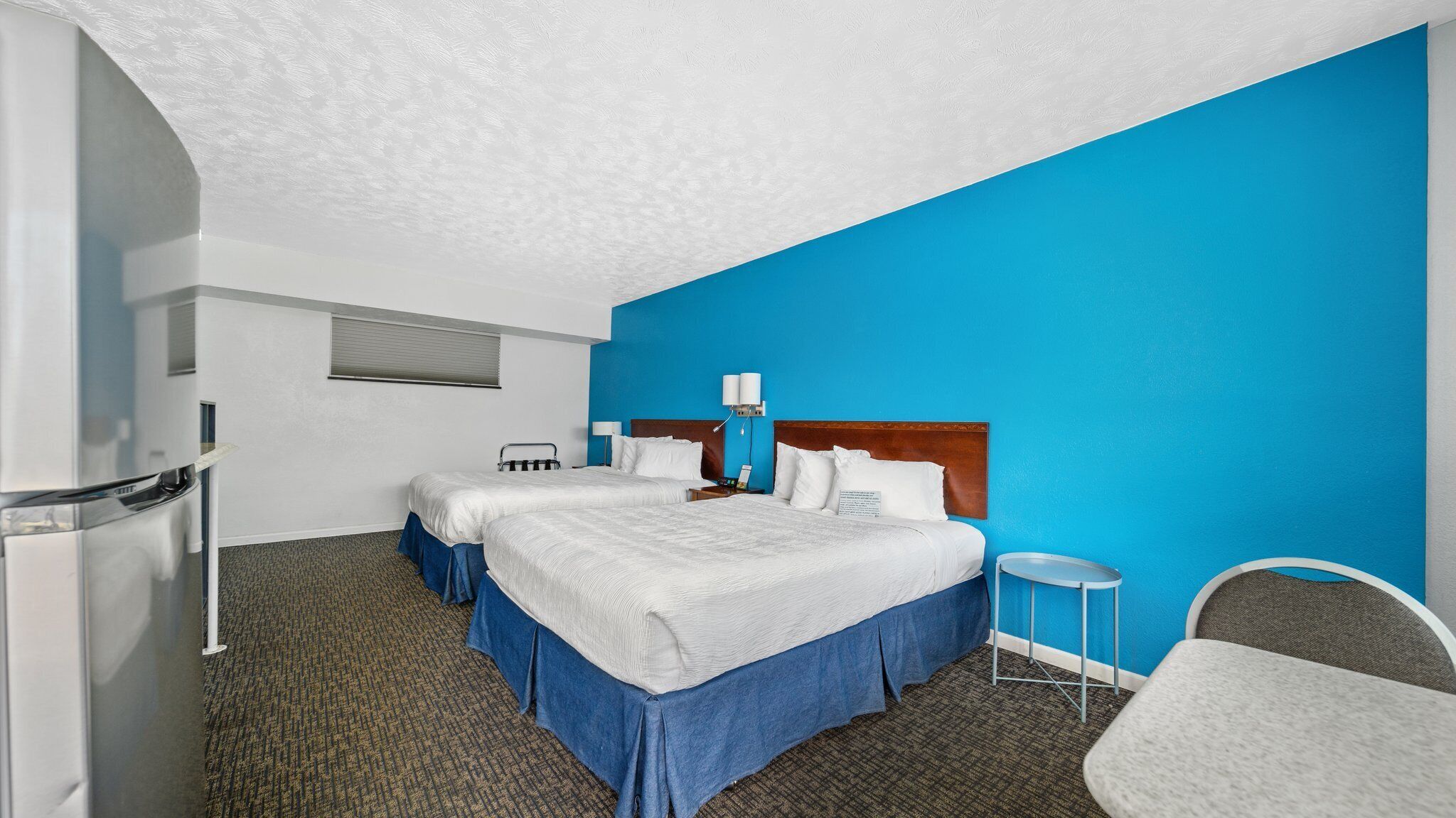A hotel room with two beds, a blue accent wall, a small round table, and a chair.