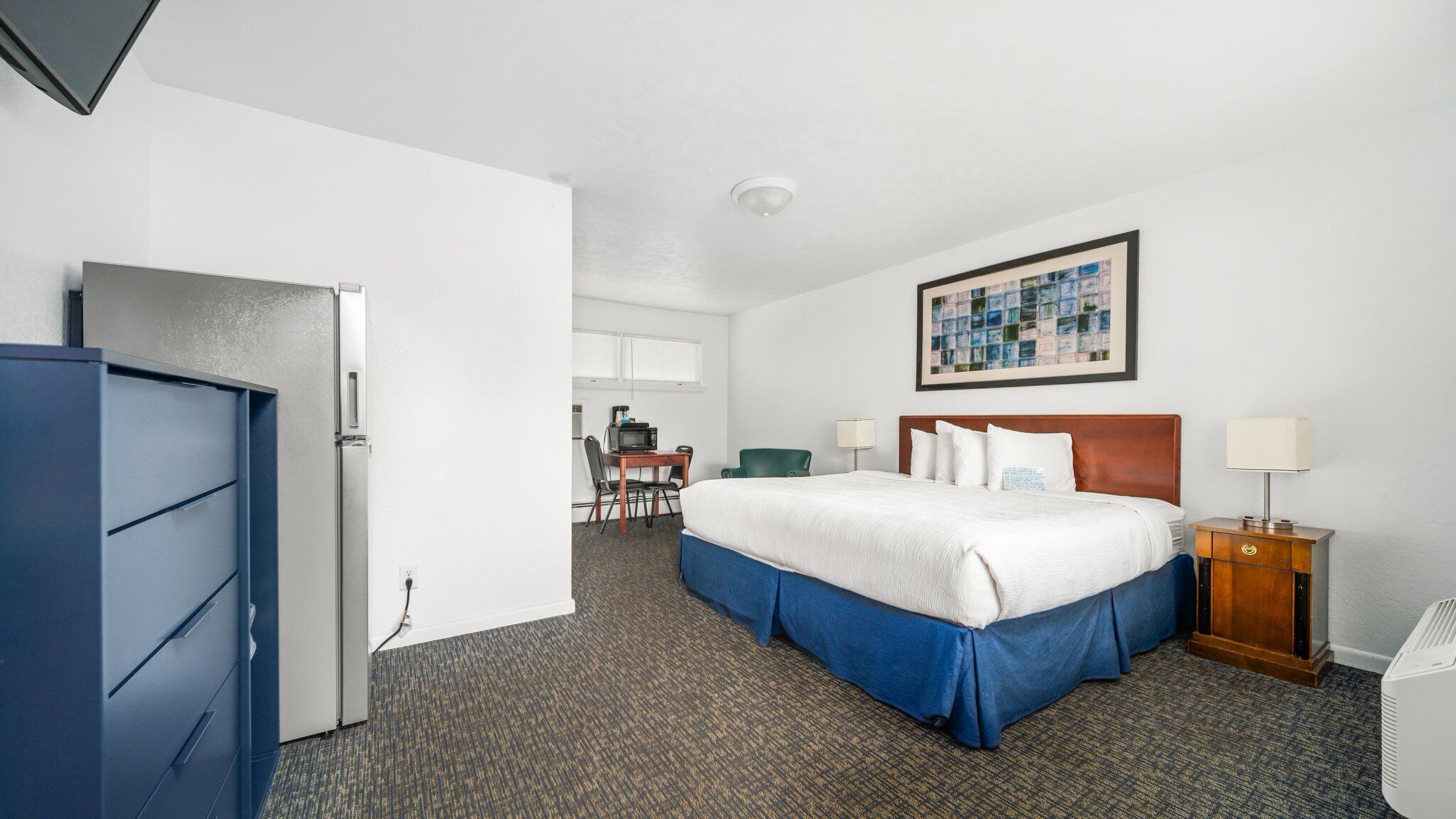 A hotel room with a bed, a desk with a chair, a microwave, a small table, a lamp, and a picture on the wall.