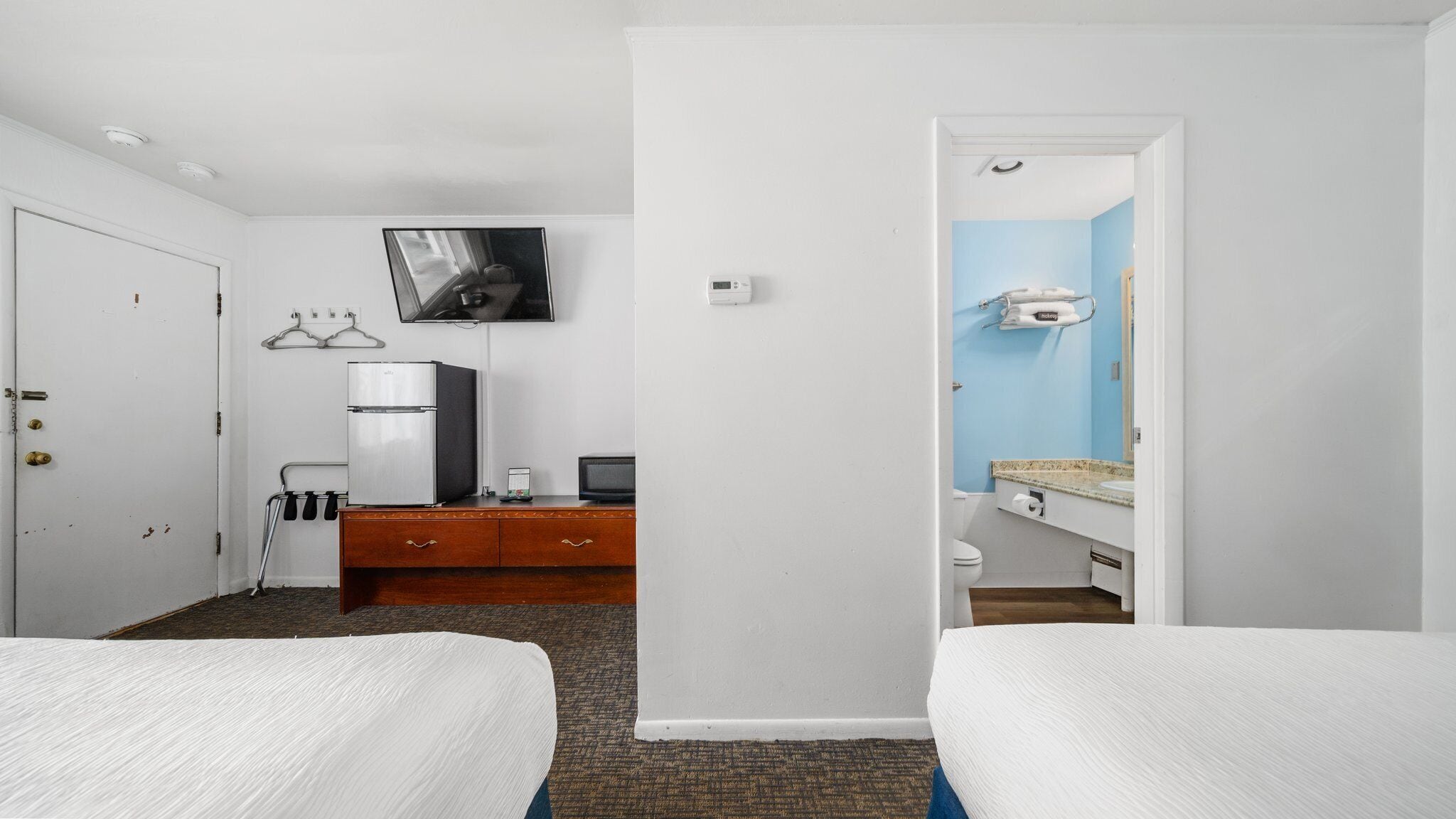 A hotel room with two beds, a desk with a computer, a TV mounted on the wall, and a bathroom visible through an open door.