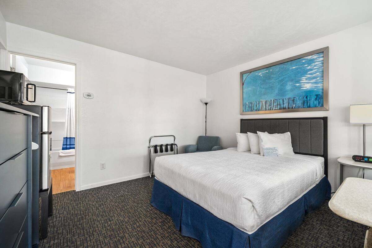 A hotel room with a bed, a desk, a chair, a wardrobe, and a painting on the wall.
