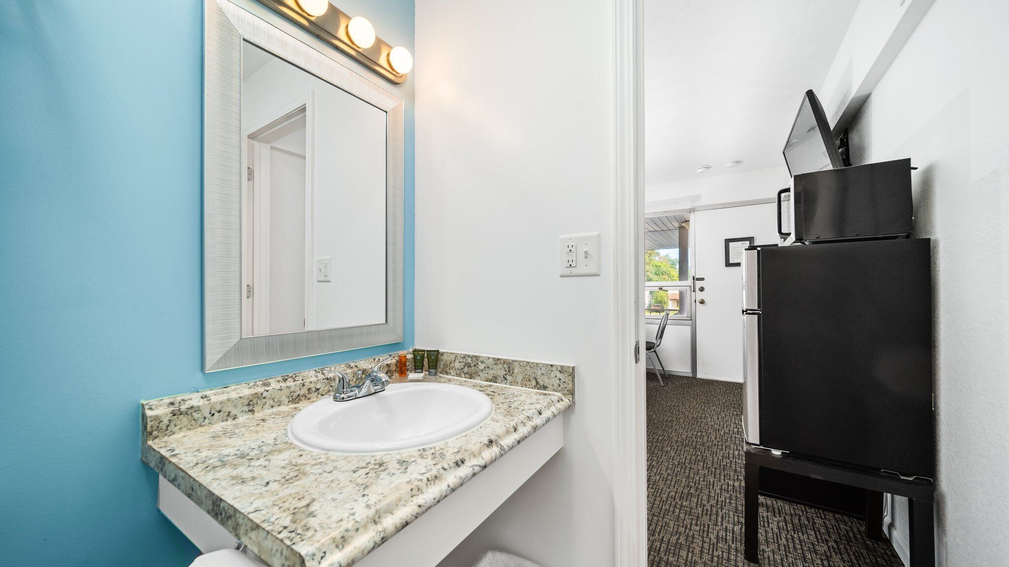A bathroom with a white sink, a mirror with lights, and a black refrigerator.