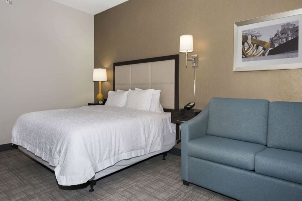 Una habitación de hotel con una cama, un sofá azul, una mesita de noche con una lámpara y un cuadro en la pared.