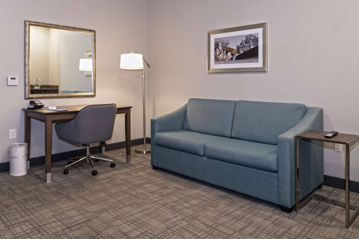 A hotel room with a desk, chair, sofa, lamp, framed picture, and a trash bin.
